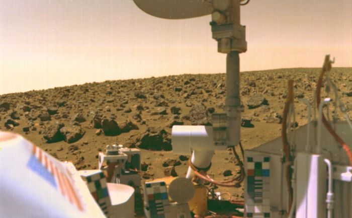 Rover’s-eye view of an orange-brown, barren landscape covered in boulders of many sizes. In the near foreground, part of the rover is captured in its own camera’s field of view.
