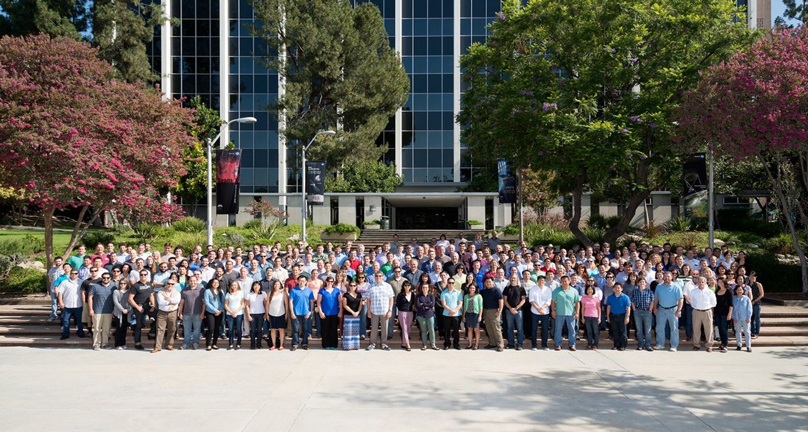 About 100 team members stand in a group photo in front of the steps of a large administration building.