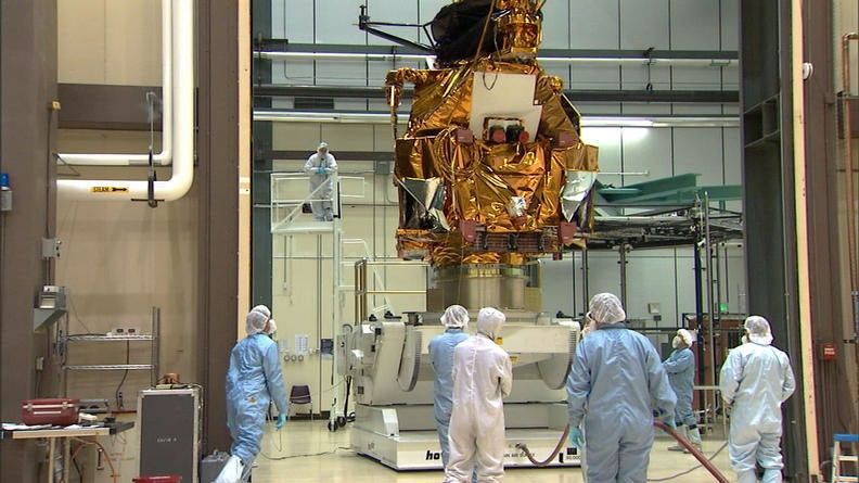 A group of eight engineers and technicians dressed in white and blue cleanroom coveralls and bonnets (called 'bunny suits') stand and watch as the large, boxy bus of the Mars Reconnaissance Orbiter is lifted by a large, white mechanism.  The bus is covered in protective, gold thermal blanketing.