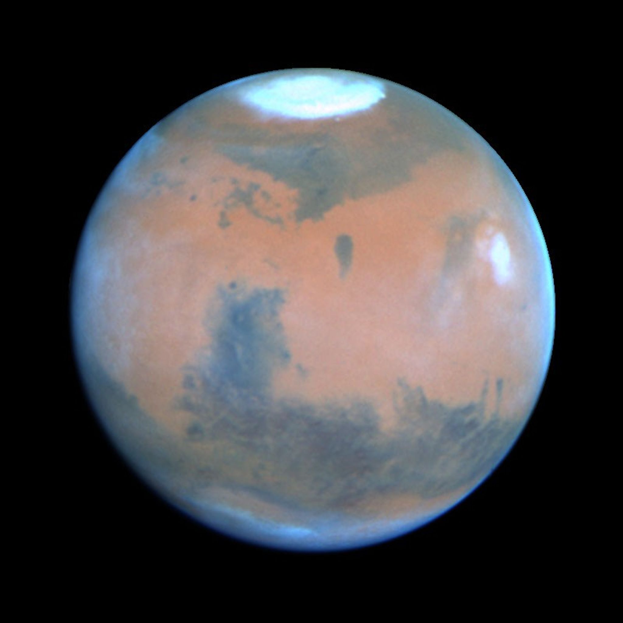 Color image of Mars taken from space, showing the planet’s reddish surface, dark volcanic regions, and bright white polar ice cap at the top.