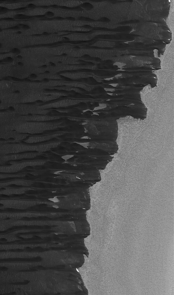 NASA's Mars Global Surveyor shows dark sand dunes in the north polar region of Mars. They are streaming away (toward the left) from a low escarpment at the edge of an outlier of polar water ice.