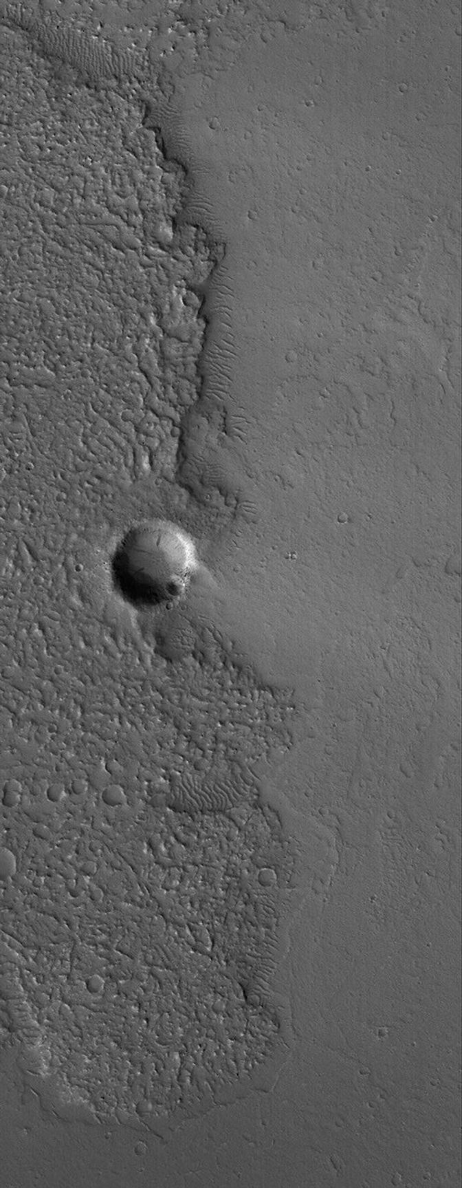 This NASA Mars Global Surveyor image shows the margin of an ancient, cratered, hummocky (rough) lava flow at just the point where it encroached upon a small impact crater east of the volcano, Tharsis Tholus.