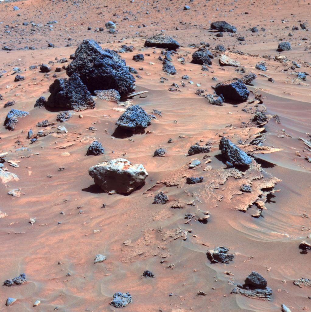 This false-color image shows paper-thin layers of light-toned, jagged-edged rocks; a light gray rock with smooth, rounded edges atop and drifts; and several dark gray to black, angular rocks with vesicles typical of hardened lava scattered across the sand