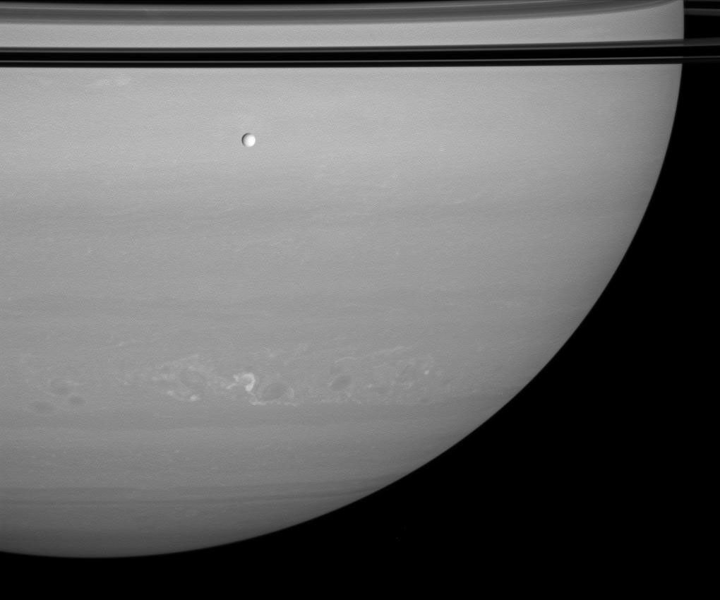 Tethys passes silently between Saturn and NASA's Cassini spacecraft as a train of storms rumbles through the planet's southern hemisphere. The rings' shadows darken the planet at top.
