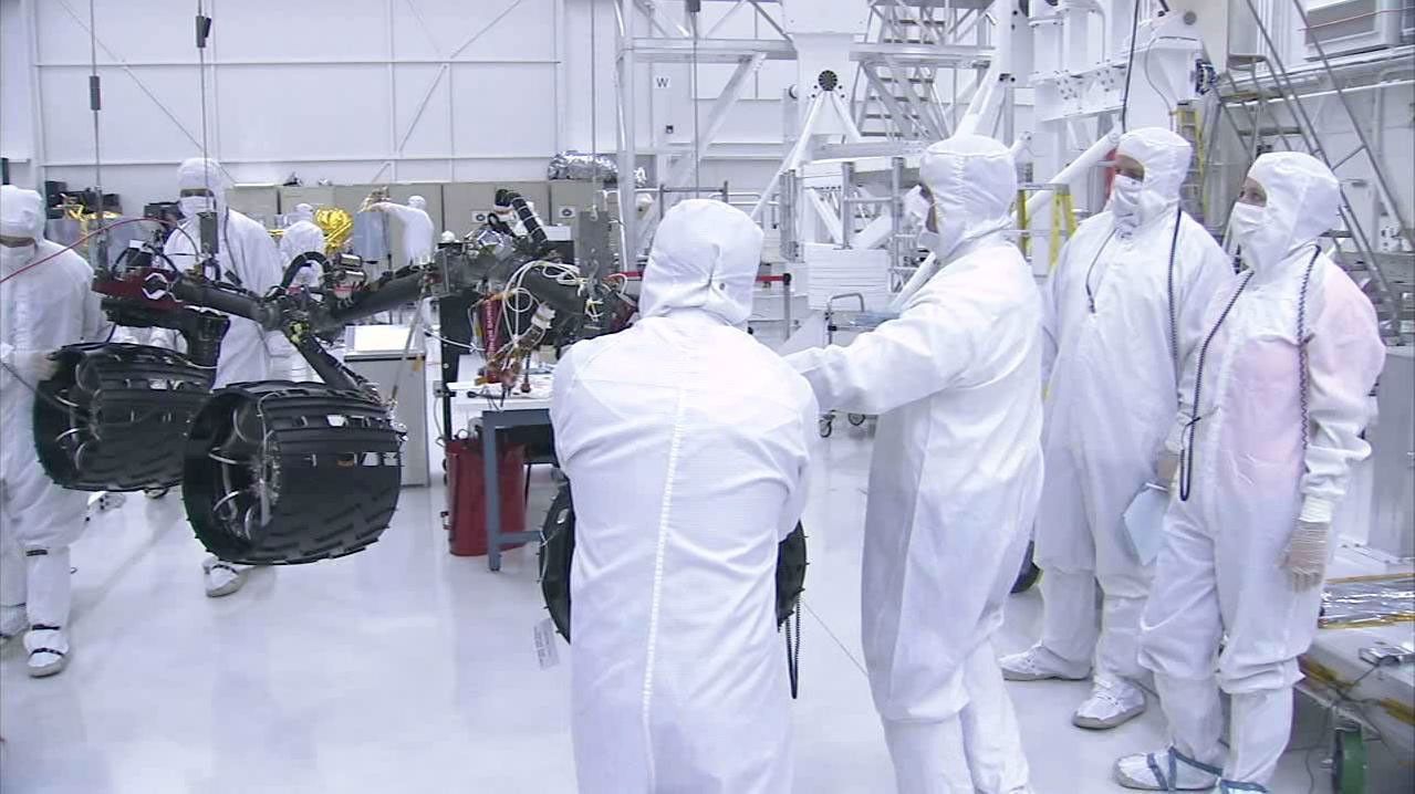 In this image taken from a video clip, NASA's Curiosity rover's wheels are shown hoisted on a pink crane and moving closer to the rover's body. Video is available at the Photojournal.