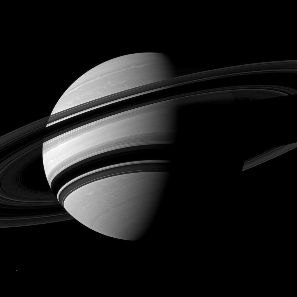 NASA's Cassini spacecraft takes an angled view toward Saturn, showing the southern reaches of the planet with the rings on a dramatic diagonal. The moon Enceladus appears as a small, bright speck in the lower left of the image.