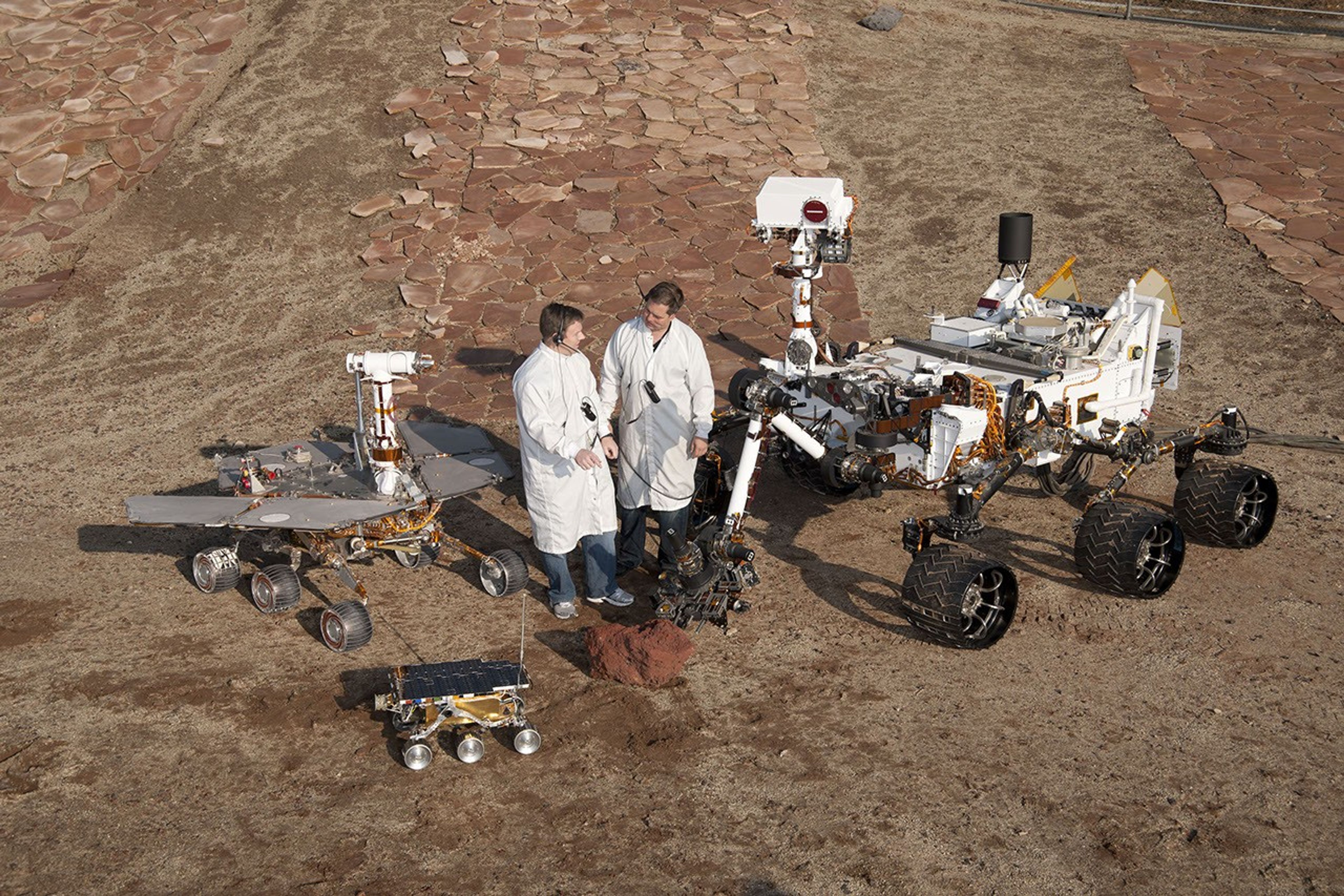 Two spacecraft engineers stand with three generations of Mars rovers developed at NASA's JPL, Pasadena, Ca. Front and center is a flight spare of Sojourner, left is a working sibling to Spirit and Opportunity, right is test rover Curiosity.