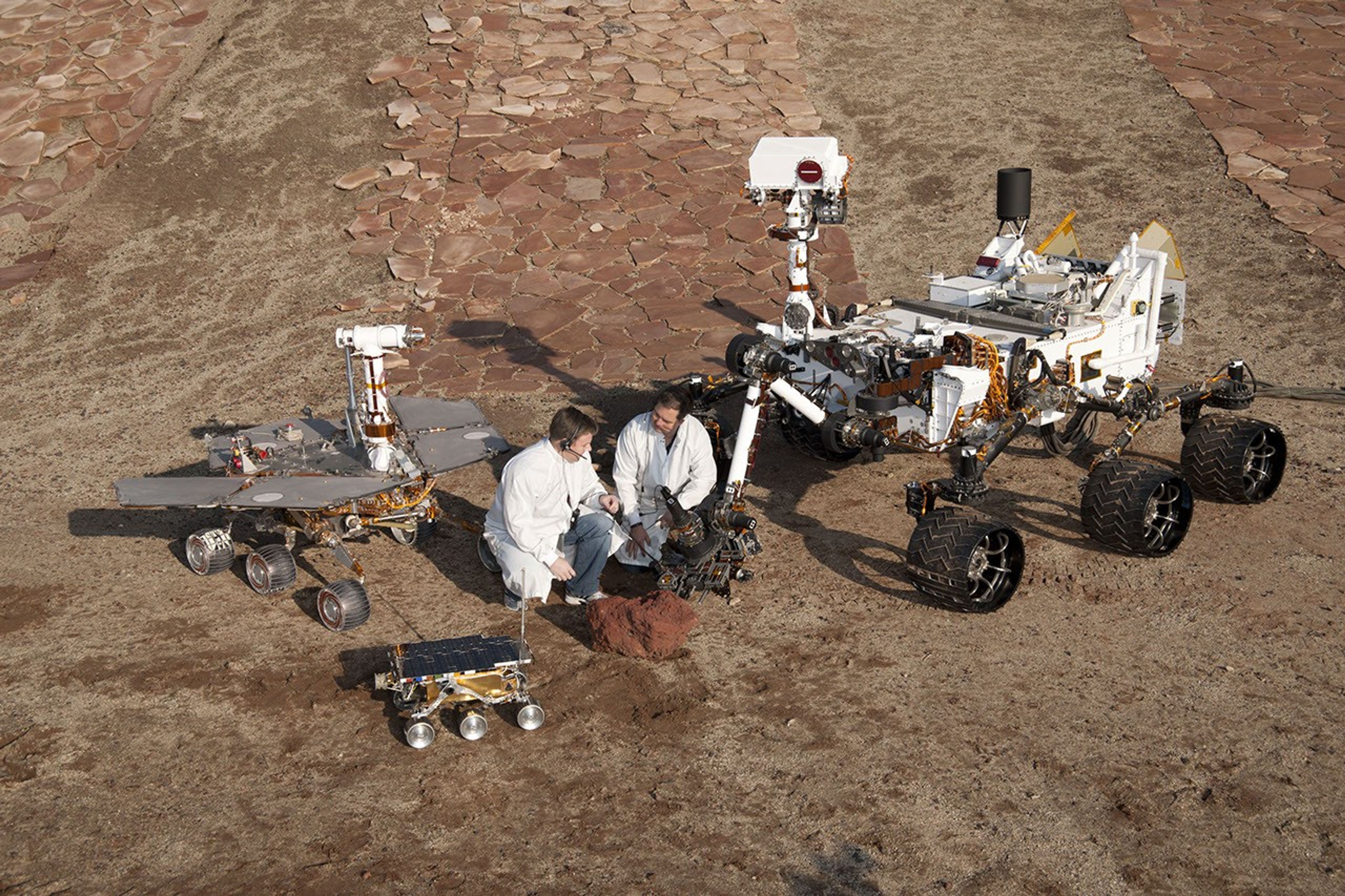 Two spacecraft engineers stand with three generations of Mars rovers developed at NASA's JPL, Pasadena, Ca. Front and center is a flight spare of Sojourner, left is a working sibling to Spirit and Opportunity, right is test rover Curiosity.