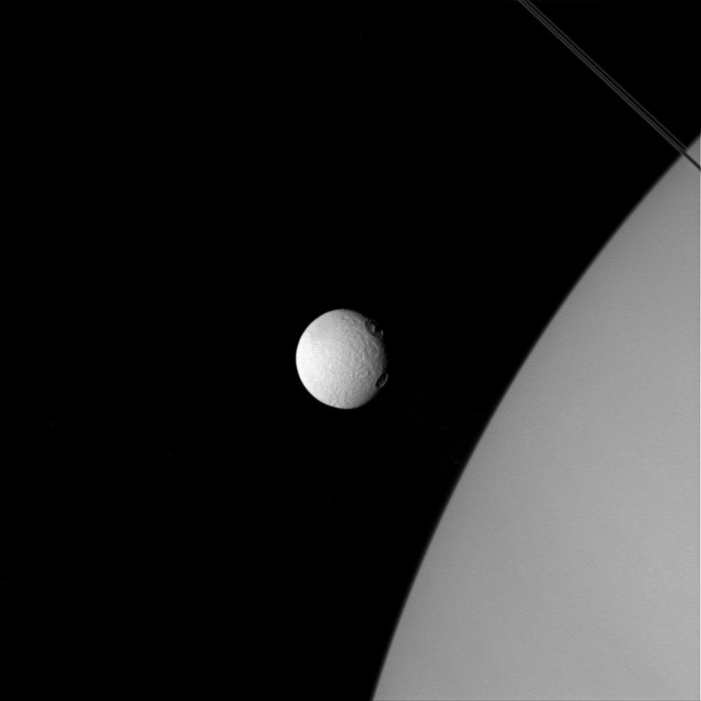 In this image from NASA's Cassini spacecraft, two large craters on Tethys, near the line where day fades to night, almost resemble two giant eyes observing Saturn. Tethys is significantly closer to the camera, while the planet is in the background.