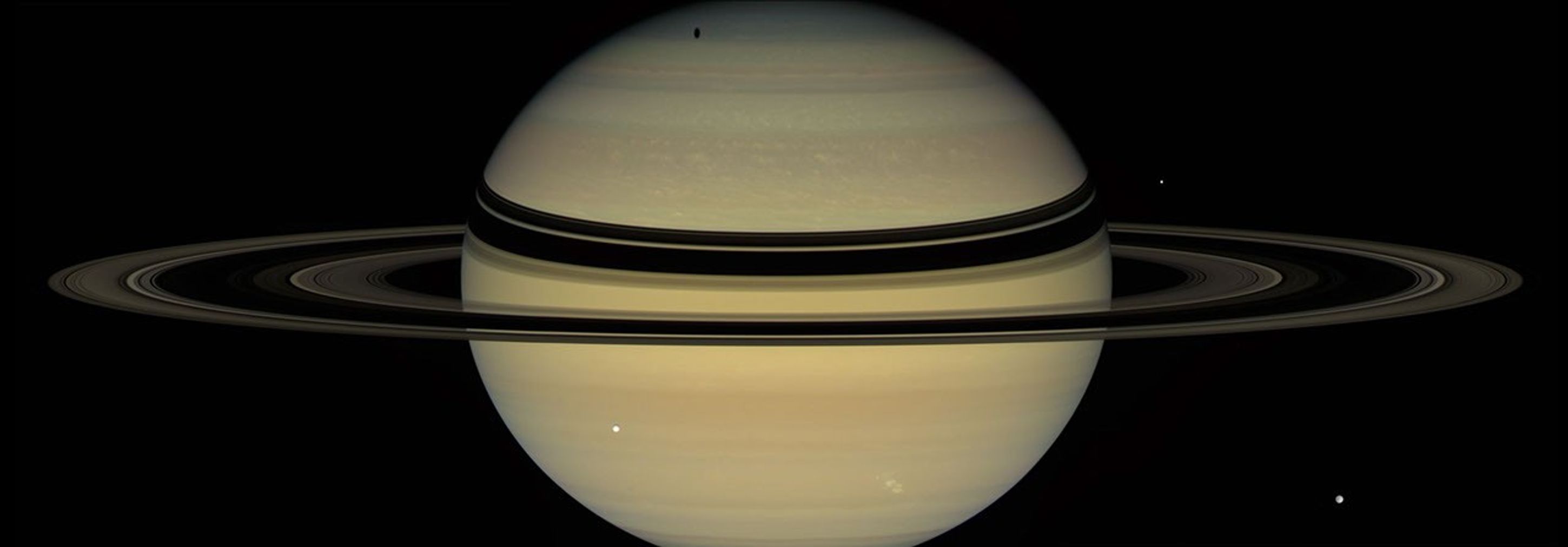 Saturn's rings cast dark bands across cloud tops in the northern hemisphere. Near the pole, an elongated shadow can be seen from Saturn's moon Tethys. Icy moons Dione (front right) and Enceladus (back right) are also seen by NASA's Cassini spacecraft.