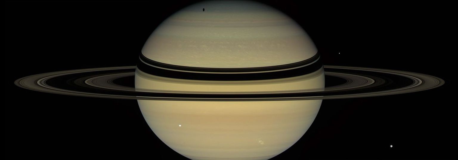 Saturn's rings cast dark bands across cloud tops in the northern hemisphere. Near the pole, an elongated shadow can be seen from Saturn's moon Tethys. Icy moons Dione (front right) and Enceladus (back right) are also seen by NASA's Cassini spacecraft.