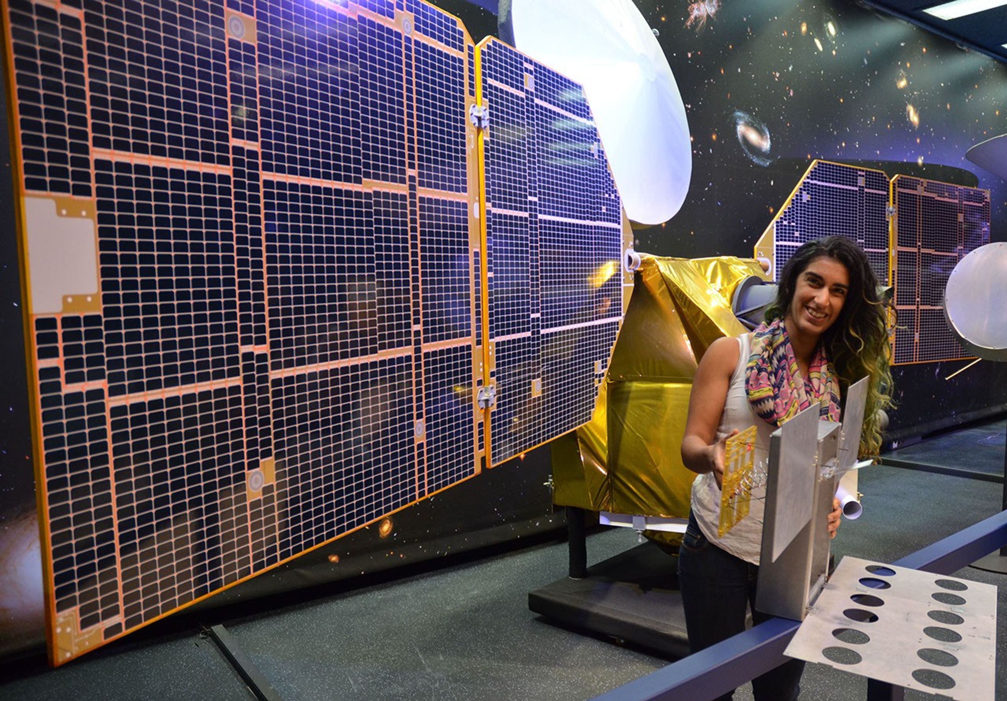 The full-scale mock-up of NASA's MarCO spacecraft held by Farah Alibay, a systems engineer for the project, is dwarfed by the one-half-scale model of NASA's Mars Reconnaissance Orbiter behind her.