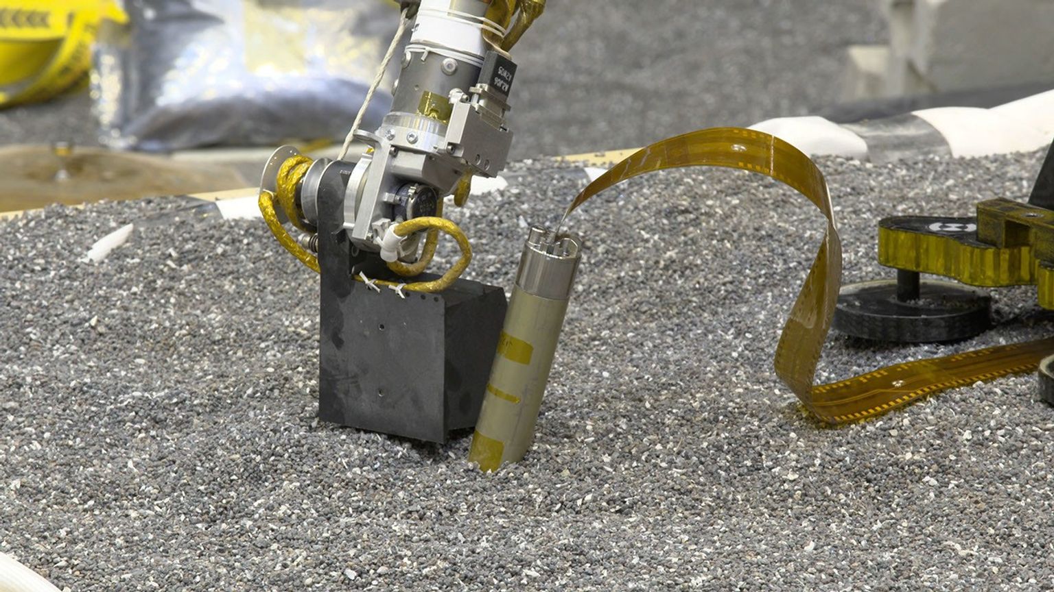 In a JPL lab, a replica of NASA InSight's robotic arm presses with its scoop on crushed garnet near a replica of the spacecraft's self-hammering 'mole.'