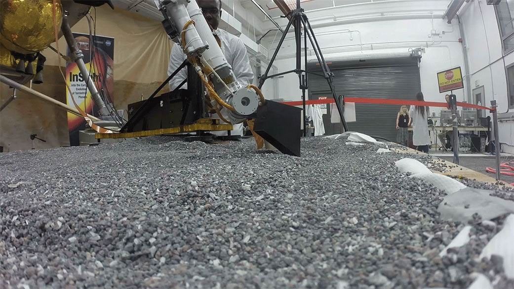 This footage from Aug. 19, 2019, shows a replica of InSight scraping soil with a scoop on the end of its robotic arm in a test lab at JPL. On Mars, InSight will scrape and tamp down soil on top of the mole to help it dig.