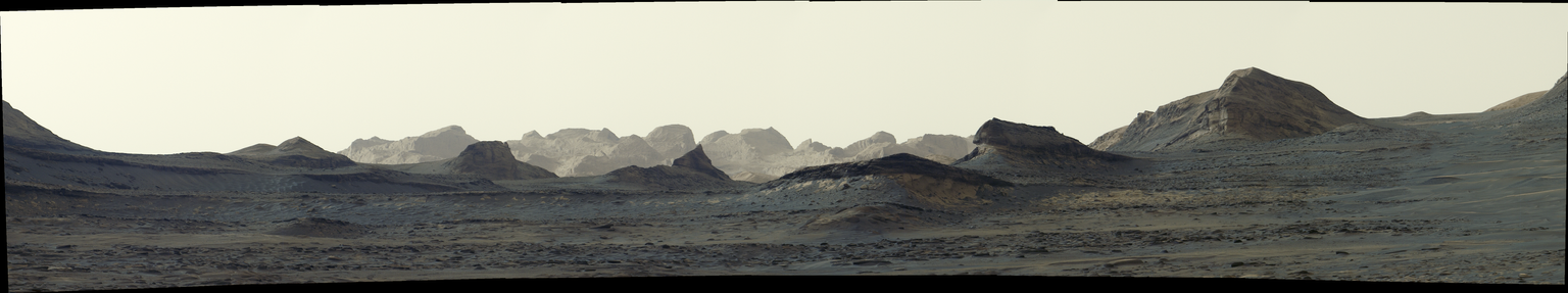 A panoramic view of a stark, grayish-tan Martian landscape featuring jagged rock formations and rolling mountain ridges beneath a pale, hazy sky.