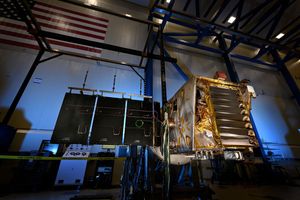 A gold spacecraft sits in a clean room with one solar panel extended.