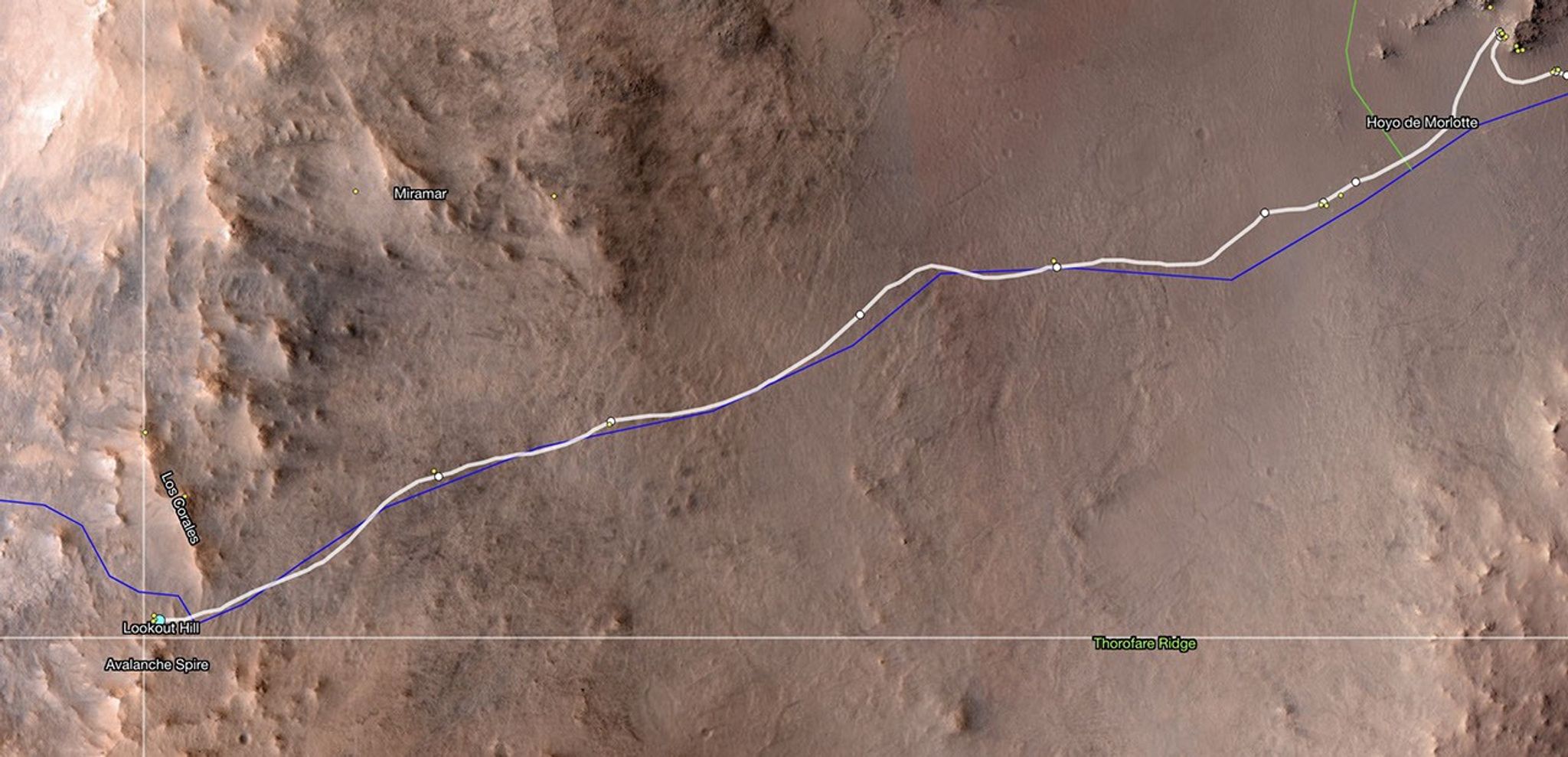 An overhead view of the Martian surface, showing the route of the Perseverance rover, marked by a weaving white line atop the landscape image. The terrain is mainly light orange-tan, with the right two-thirds mostly flat, and the left third appearing more rocky and hilly. The rover’s path runs diagonally into that section, from the upper right corner of the image to the lower left corner.