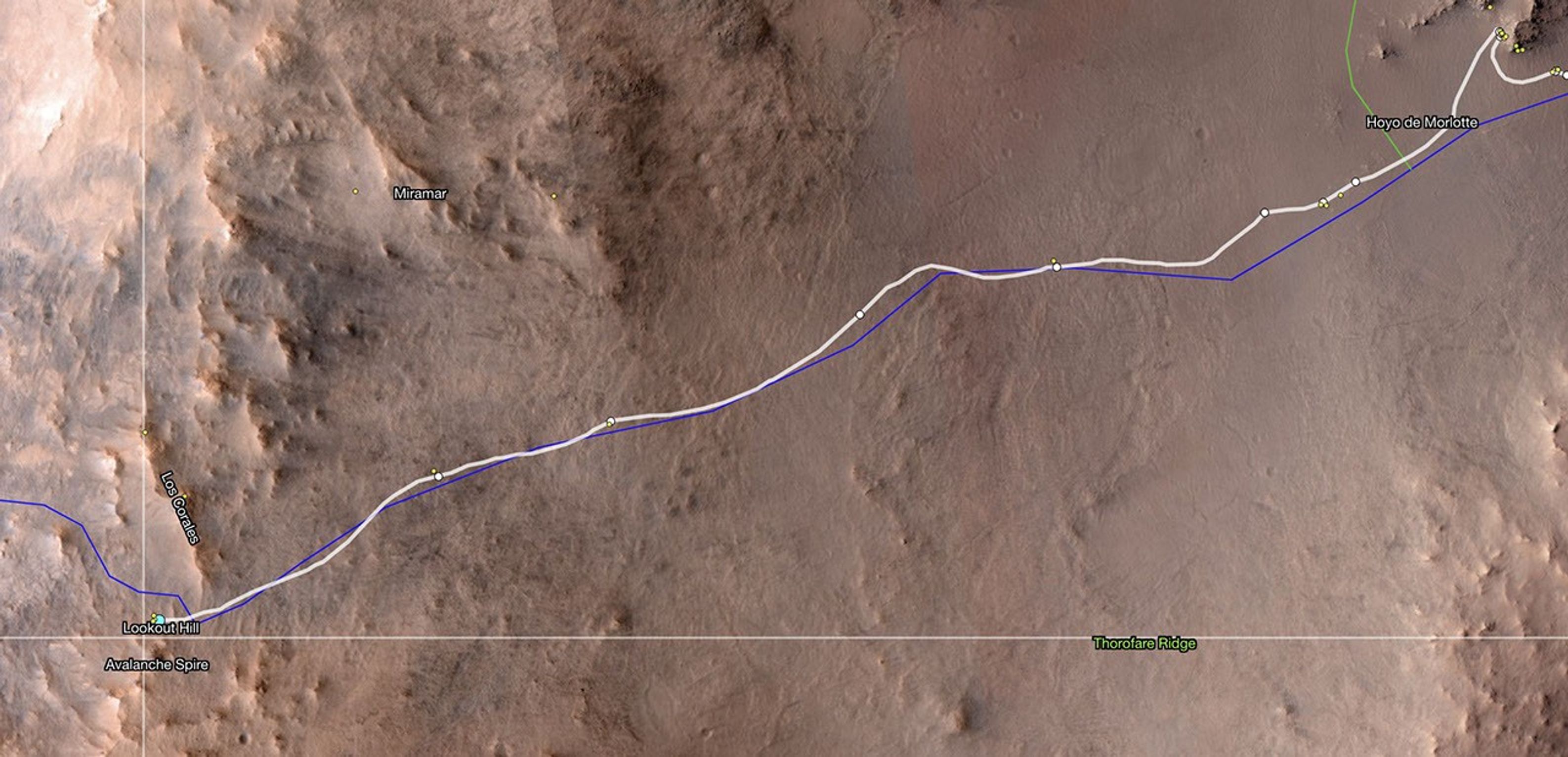 An overhead view of the Martian surface, showing the route of the Perseverance rover, marked by a weaving white line atop the landscape image. The terrain is mainly light orange-tan, with the right two-thirds mostly flat, and the left third appearing more rocky and hilly. The rover’s path runs diagonally into that section, from the upper right corner of the image to the lower left corner.