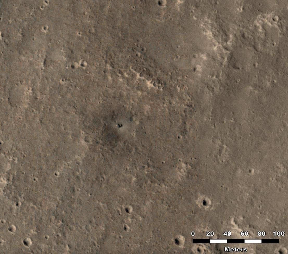 An overhead view of the Martian surface, shows dark tan terrain with scattered craters. At the center of the image is a dark spot in a depression that appears smoother and darker than the ground around it, resembling fabric that a pointed stick had dented, then punctured.