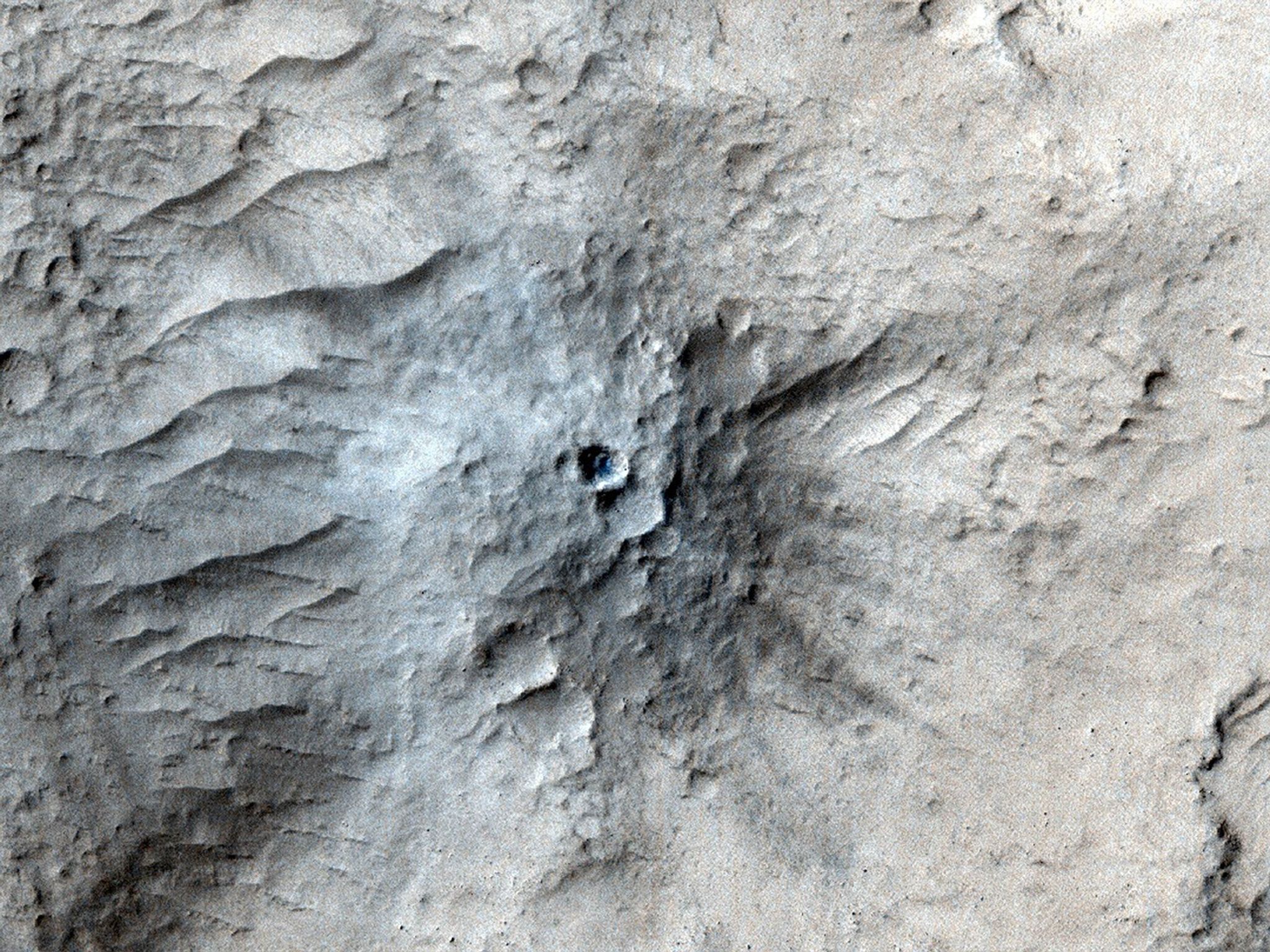 An overhead view of the surface of Mars shows the ground in shades of very light tan, with uneven, wrinkled terrain, with what looks like a mound or anthill rising from the center with a dark hole in the middle. Some areas around the center are tinged in shades of blue, while others around the edges of the image are tinged in shades of brown. Then right side of the mound appears to be shaded, and dark gray lines emanate from the hole like rays.