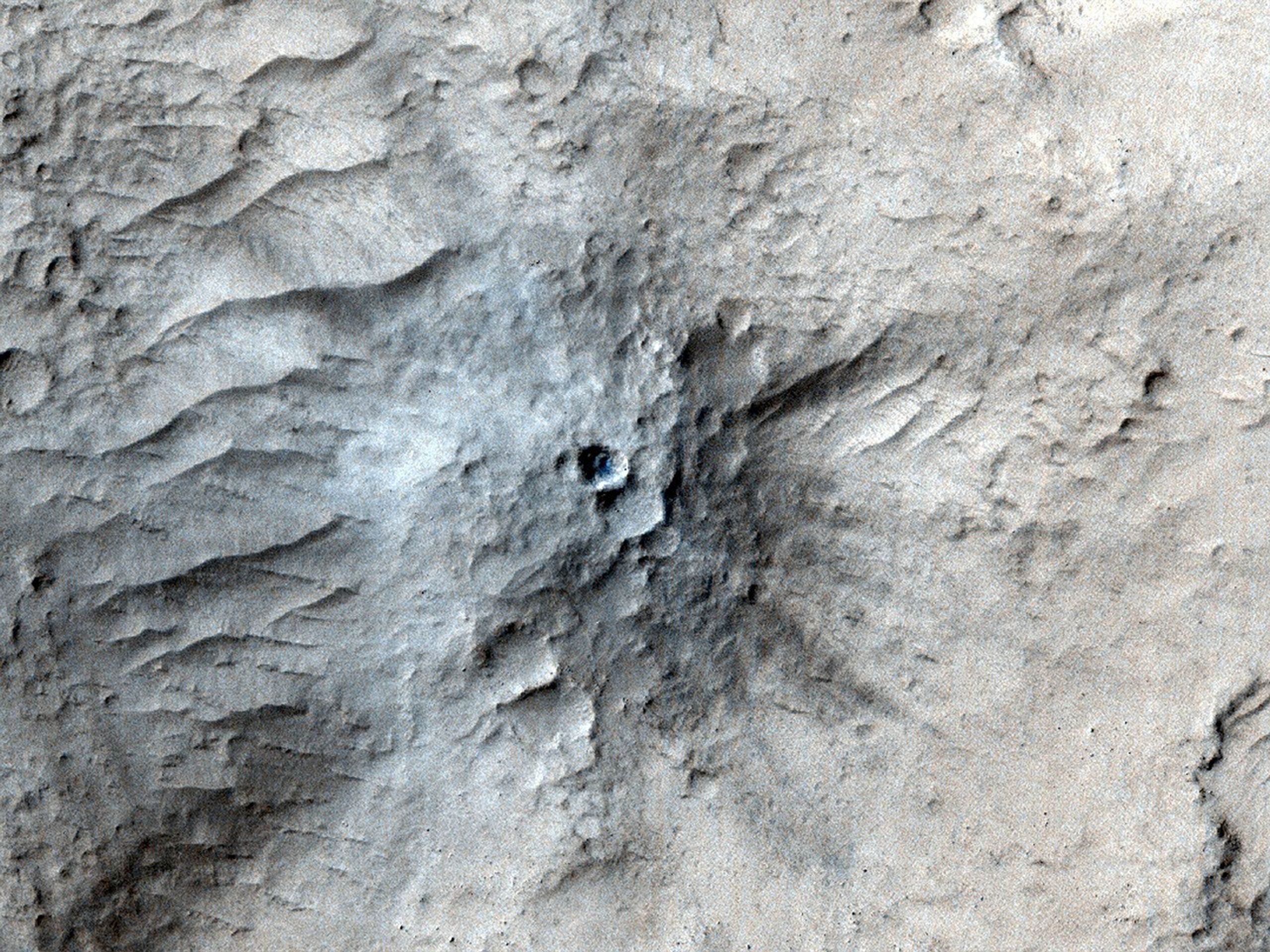 An overhead view of the surface of Mars shows the ground in shades of very light tan, with uneven, wrinkled terrain, with what looks like a mound or anthill rising from the center with a dark hole in the middle. Some areas around the center are tinged in shades of blue, while others around the edges of the image are tinged in shades of brown. Then right side of the mound appears to be shaded, and dark gray lines emanate from the hole like rays.