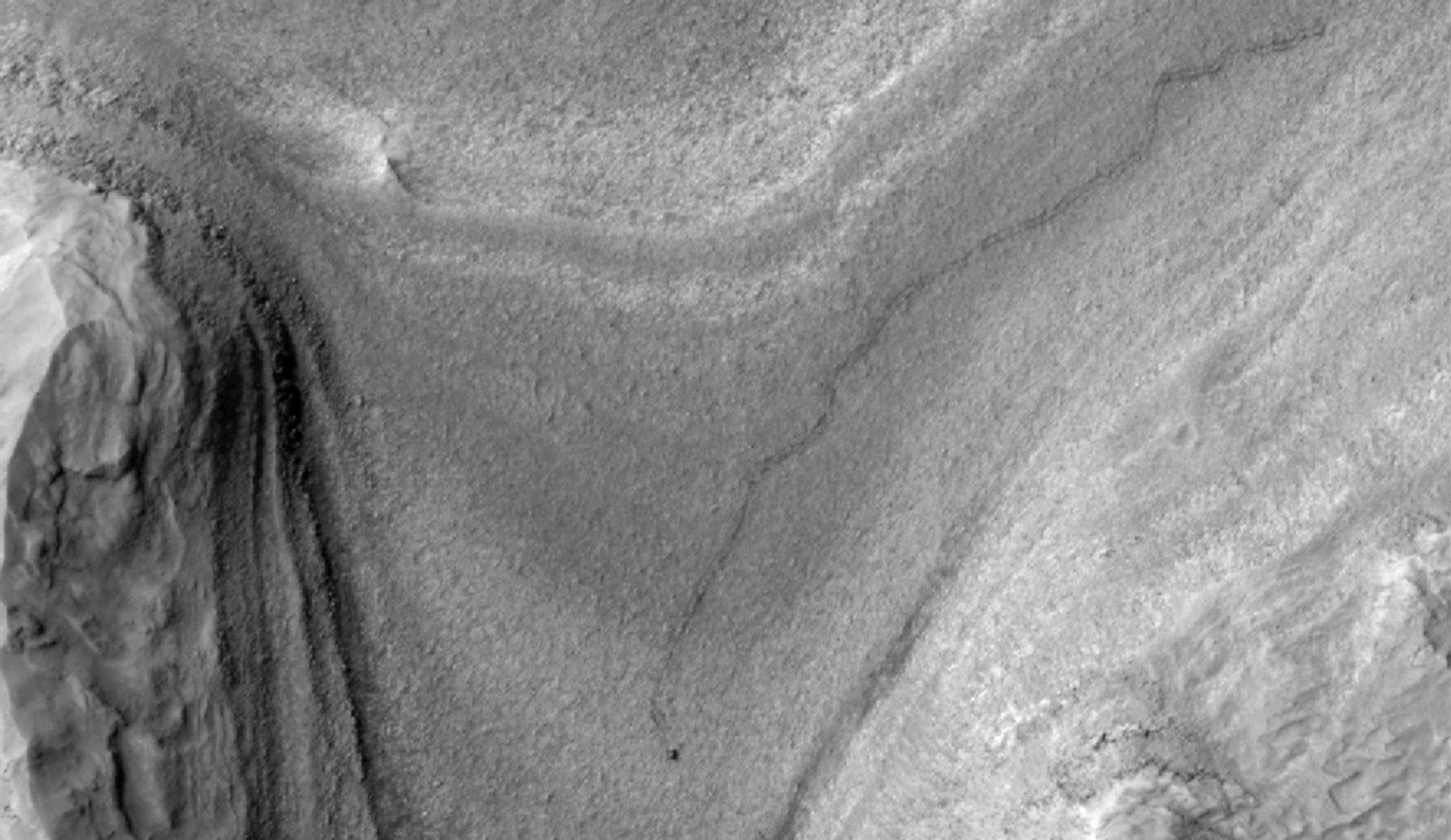 Grayscale aerial photo of the Martian surface, showing terrain far below that forms a V-shape. The left side is bordered by layered rock rising up to form a steep hill; the right side is also layered ground rising from the surface, but appears to be a gentler slope. A dark, meandering line traces on the ground from the upper right corner toward the bottom center of the image. The line ends at a black dot just above the bottom edge of the frame.