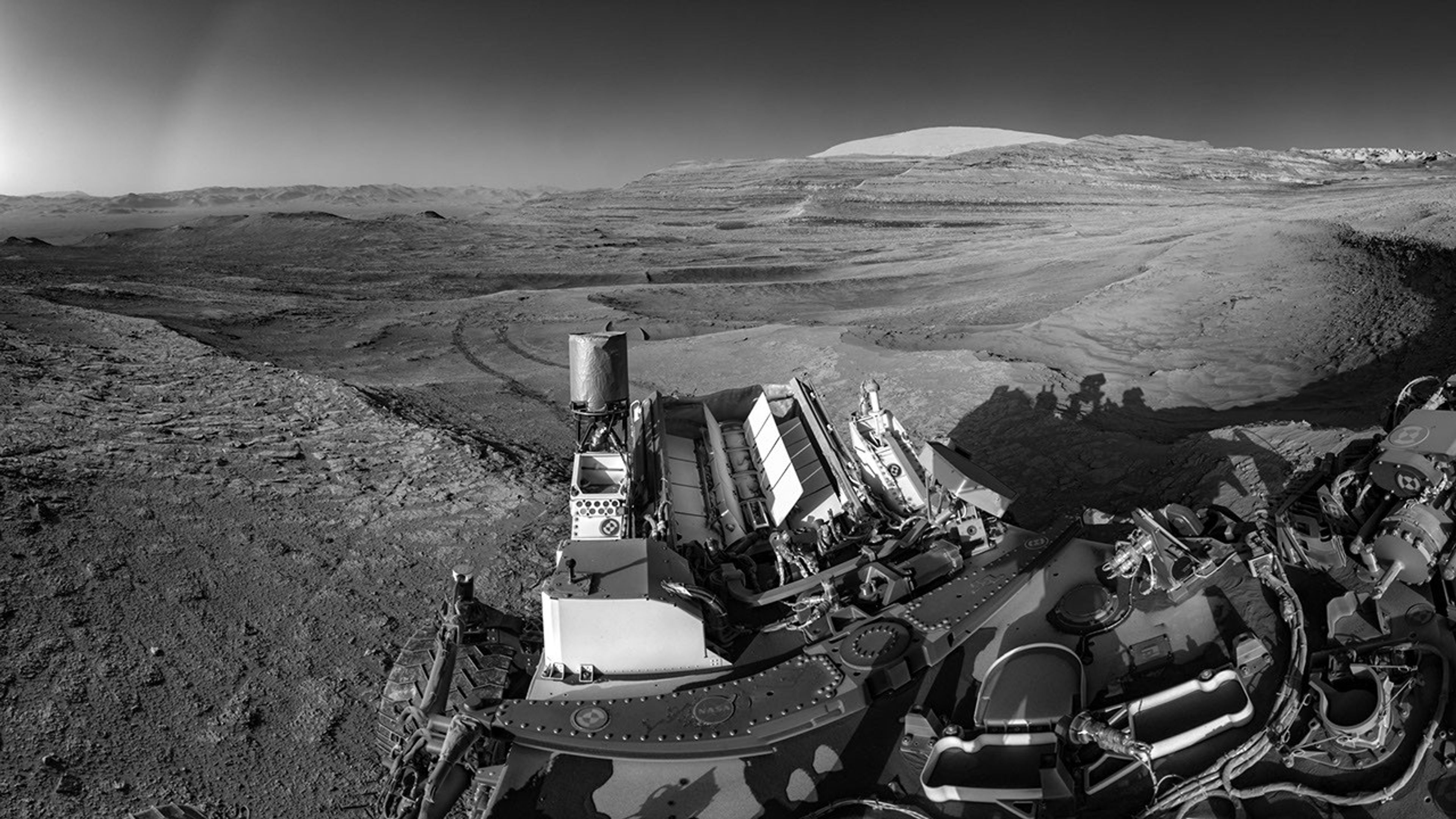 A grayscale, wide-angle landscape photograph of the Martian surface. The rocky terrain is dark gray and stretches into the distance where a whitish-gray mountain rises at the upper right -center of the image. Much of the rover is visible in the lower right corner of the image, in great detail.