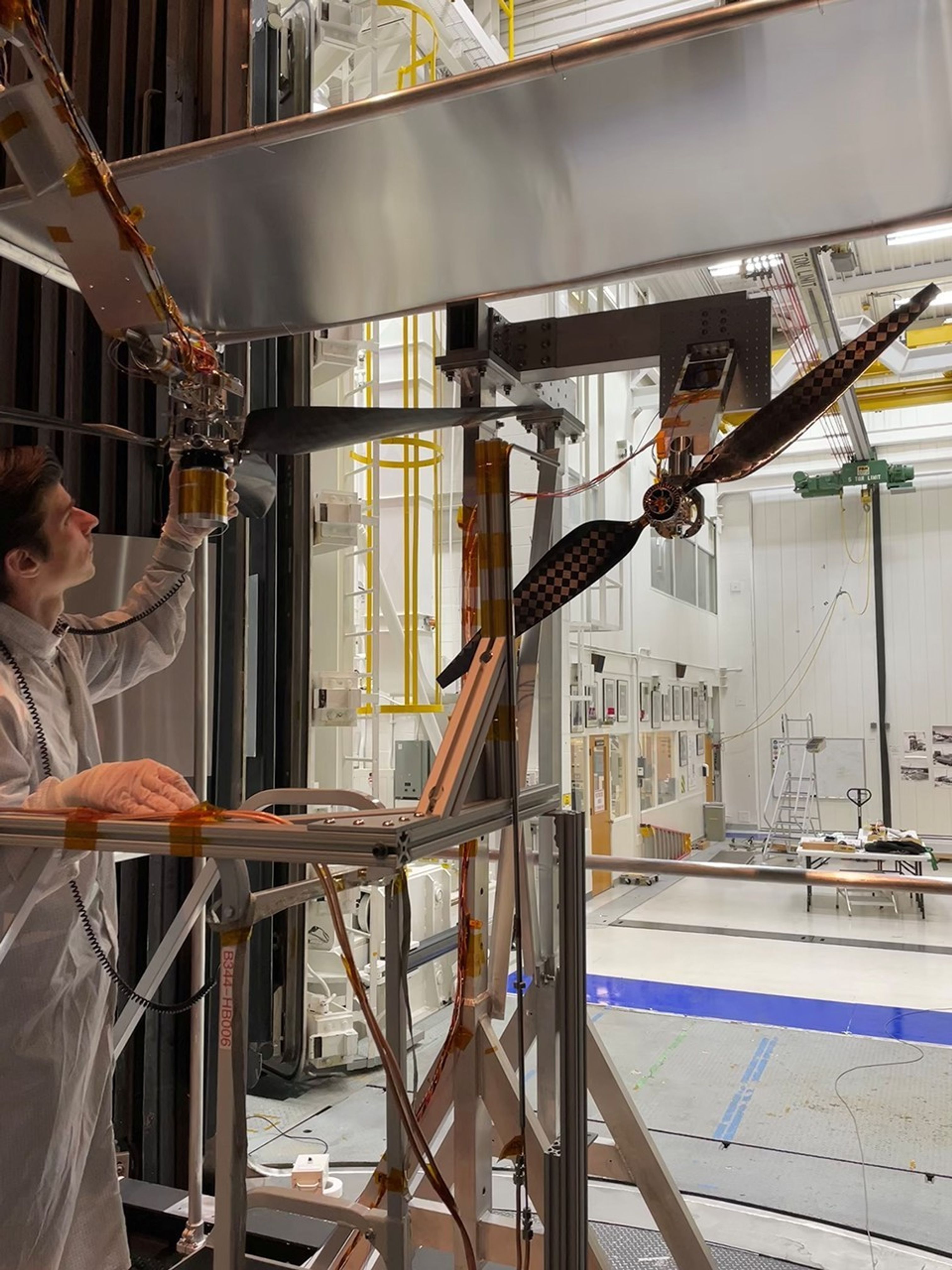 A man in a white clean room suit inspects a horizontal three-bladed rotor. To the right, a vertical two-bladed rotor with a checkered pattern is mounted. Both sit within a large, white industrial testing chamber filled with scaffolding and equipment.