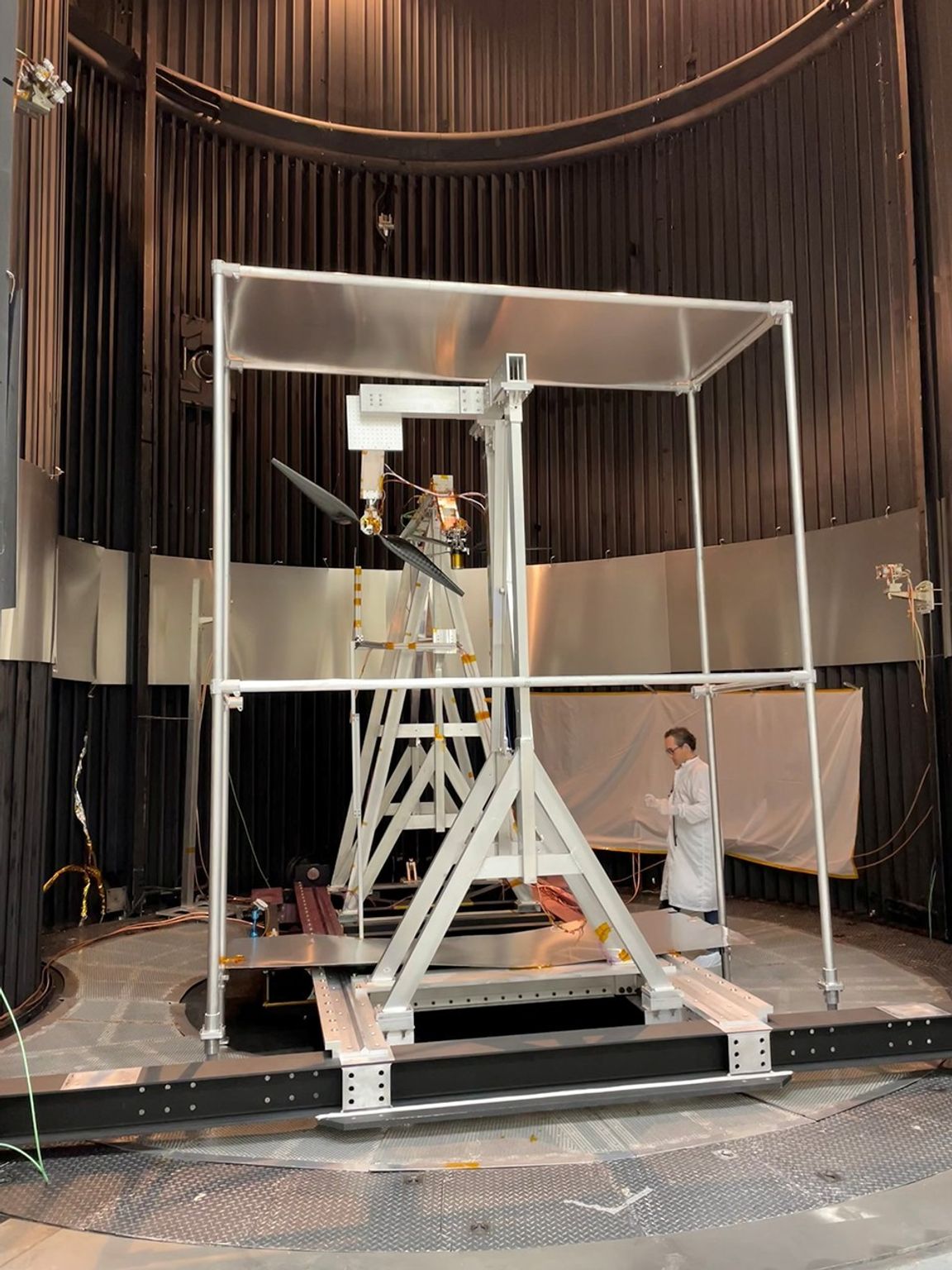 A wide shot inside a dark, cylindrical testing chamber with vertically ribbed walls. In the center, a large silver metal support structure holds a rotor with two long, dark blades. A person in a white lab coat stands to the right of the rig.