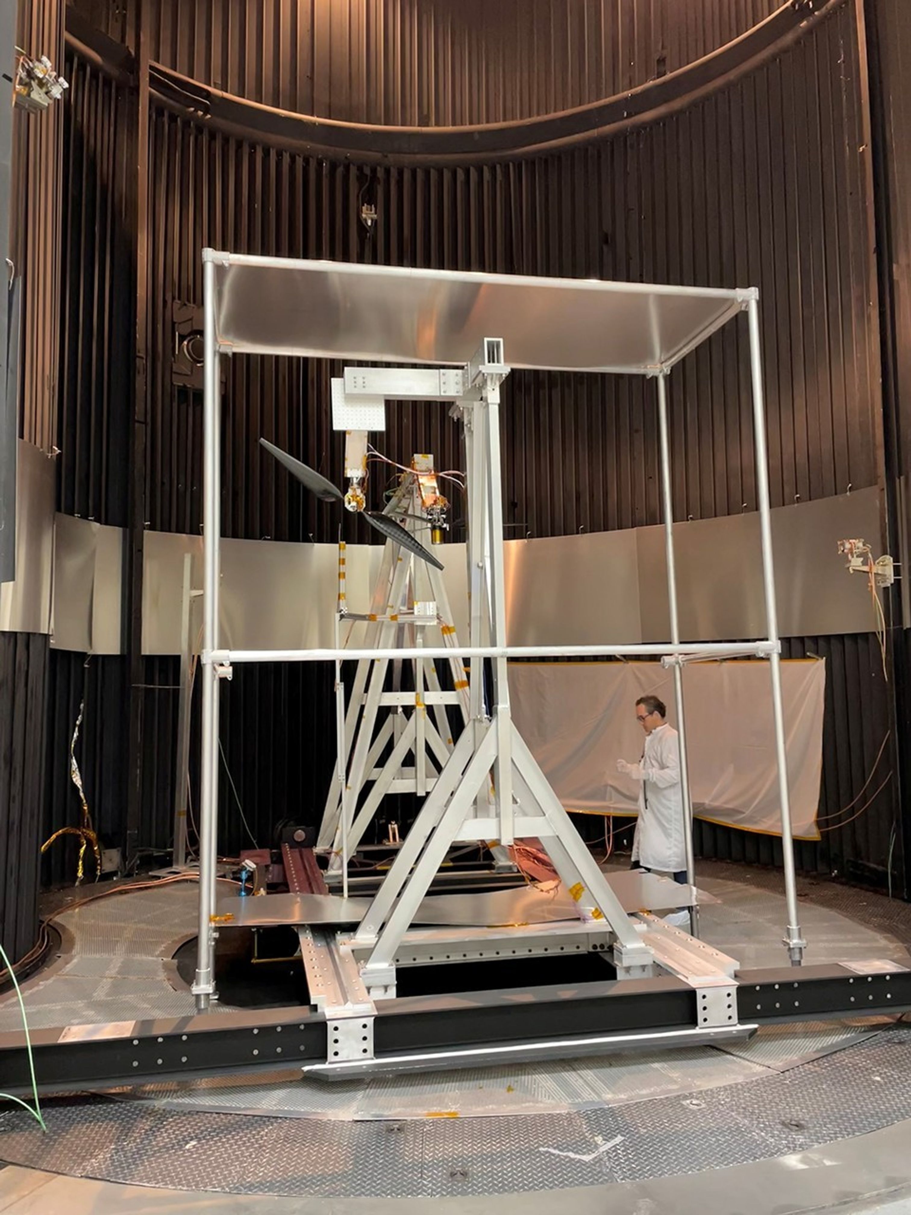 A wide shot inside a dark, cylindrical testing chamber with vertically ribbed walls. In the center, a large silver metal support structure holds a rotor with two long, dark blades. A person in a white lab coat stands to the right of the rig.