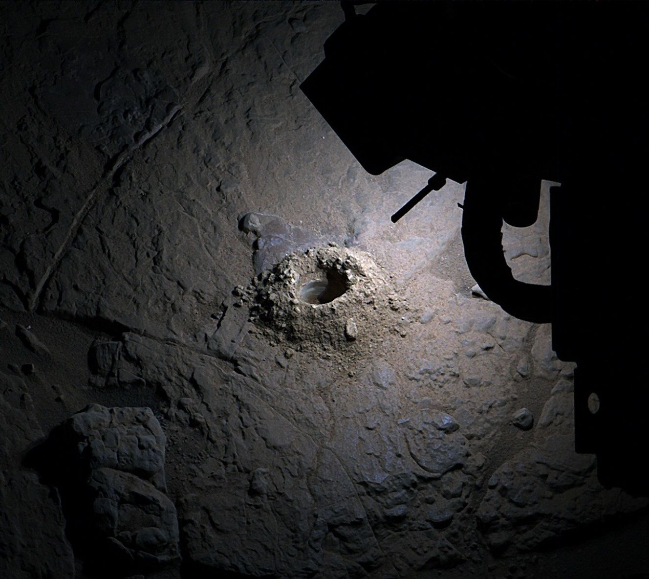 A color photograph, taken at night on the Martian surface, shows a hole drilled into a mostly flat slab of brownish-gray rock, at the center of the frame. The hole is dark, and surrounding its opening is mounded lighter-toned, tan soil and small rocks, extracted from the hole by the drill. The scene is illuminated by artificial light, but fades to darkness around the edges of the image. The upper right corner of the image is blocked by a portion of the Curiosity rover, unlit and creating a black silhouette over that part of the scene.