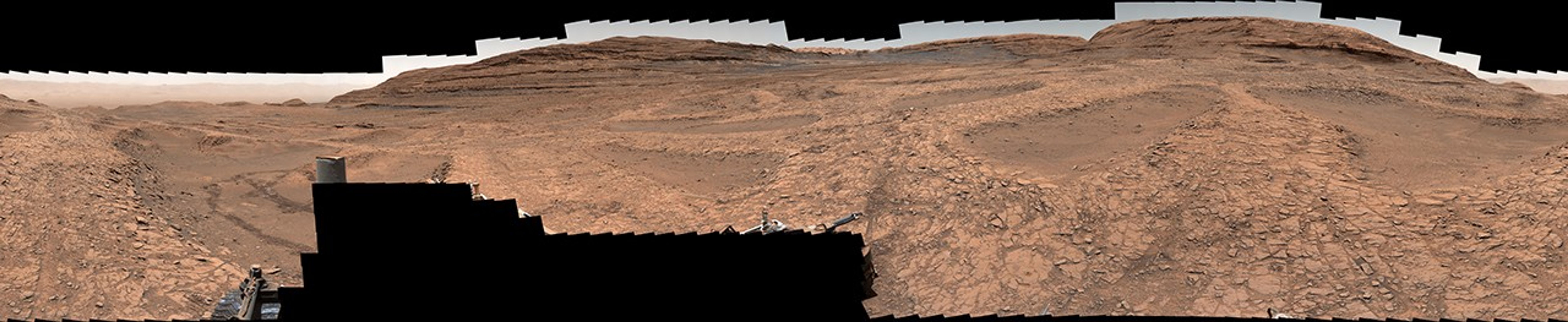 A series of shallow, sand-filled pits with low ridges spread across a tawny Martian landscape. Rover tracks stretch toward the horizon at left, and steep ridgetops loom in the background.
