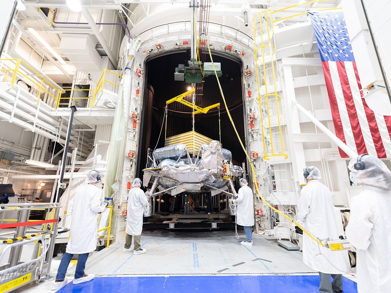 Engineers in white clean-room garb and hairnets guide a spacecraft suspended from a crane into a large thermal vacuum chamber inside a high-bay facility with an American flag hanging on the wall.