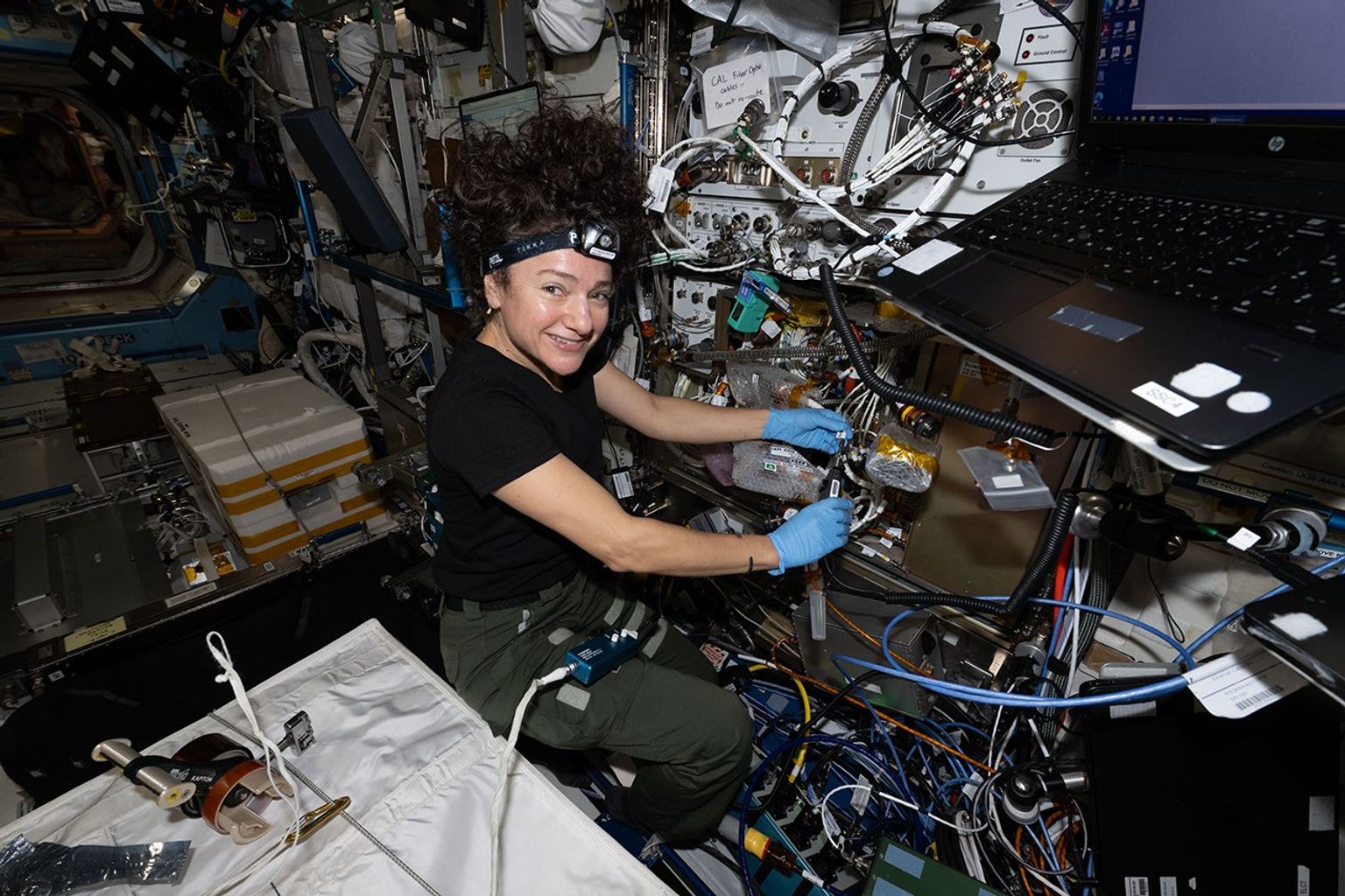 A woman in a black T-shirt with a headlamp affixed to her head and her hair floating up above her wears latex gloves to handle cabling that she’s working on. Wires, a laptop computer, and other equipment surround her.