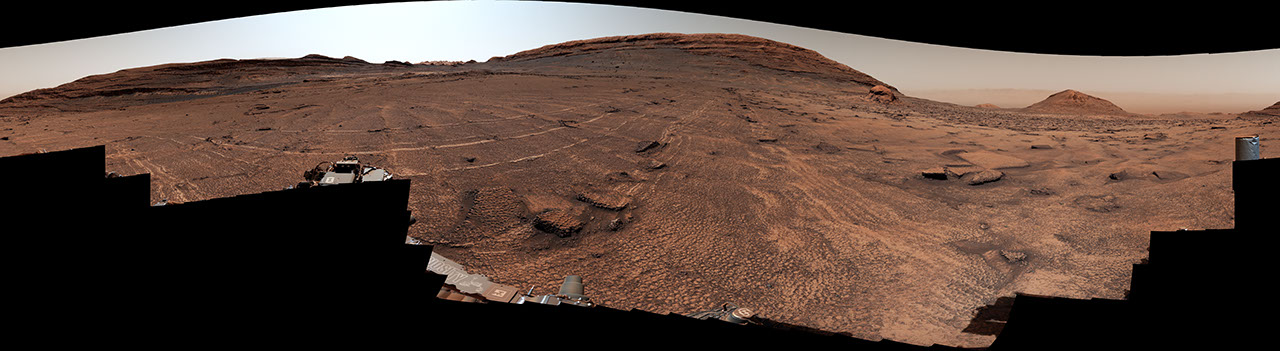 A panoramic view of a sweeping Martian plain covered in patterned fractures leading toward distant hills showcases the terrain explored by NASA's Curiosity rover to investigate the planet's past environment.