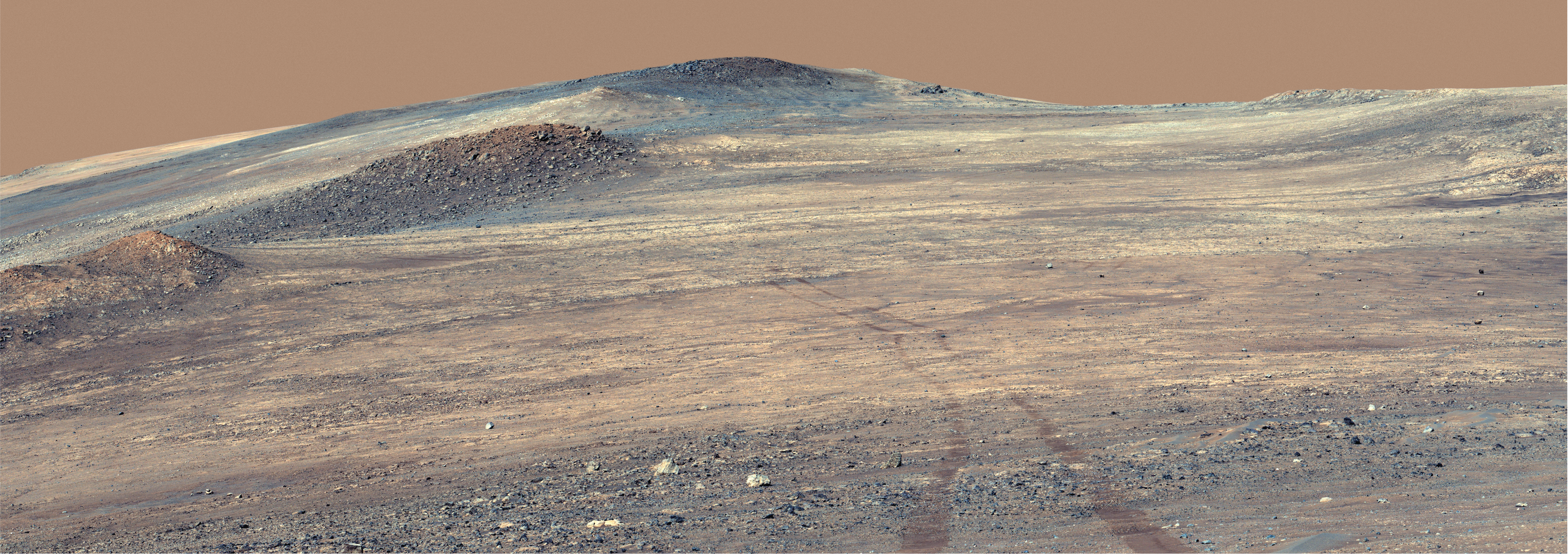 A rocky Martian hillside under a reddish sky, with distinct rover tracks leading across the foreground toward higher elevations.
