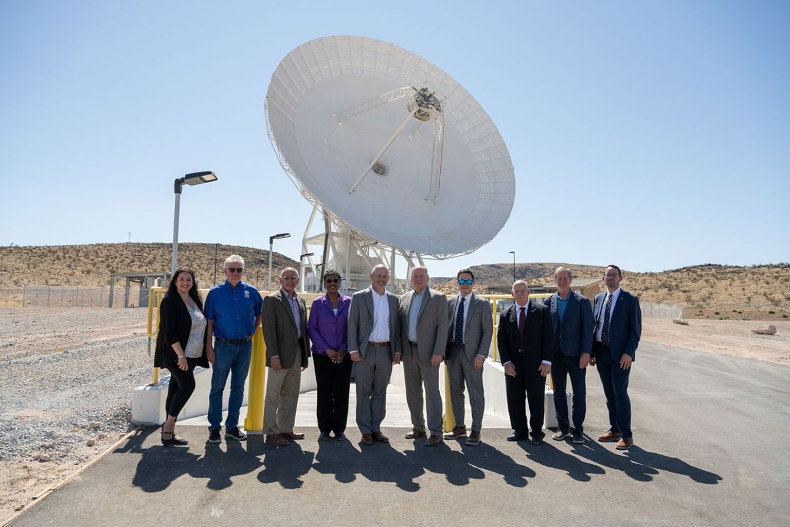 Ten people in professional attire pose together outside under a clear blue sky, with a massive white satellite dish standing directly behind them.