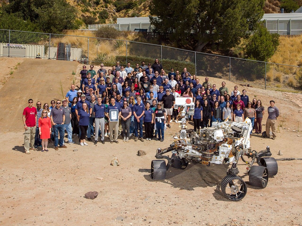 About 100 members of NASA’s Perseverance Mars rover mission team stand posed together in a sandy outdoor test facility alongside a full-scale Mars rover model.