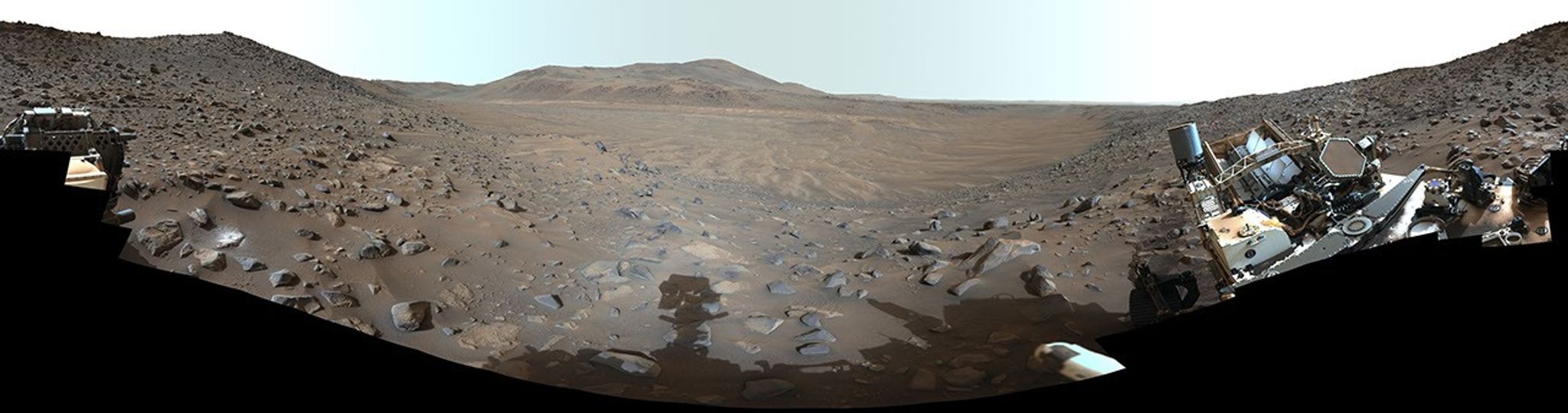 Una vista panorámica de un paisaje árido y rocoso con colinas a lo lejos, enmarcada por partes de un rover robótico visibles en los bordes izquierdo y derecho.