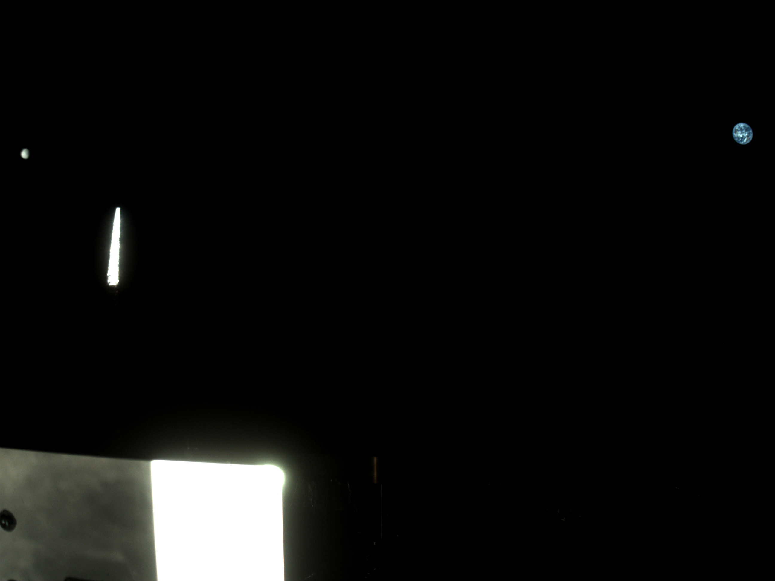 The Moon and Earth are two tiny dots on opposite sides of the black void of space. The Moon is a tiny white dot on the far left side of the image. Earth is a slightly larger dot made of shades of swirling blue on the right. In the foreground, bright polygons of light reflect back at the imager in white.