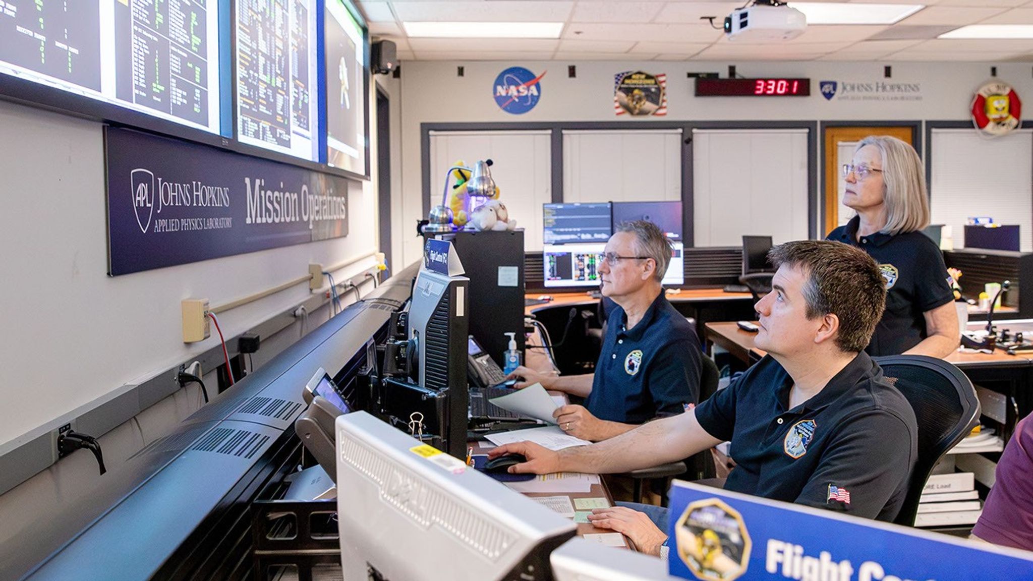 Flight operators at computers in a mission control center monitor spacecraft data on large wall displays.