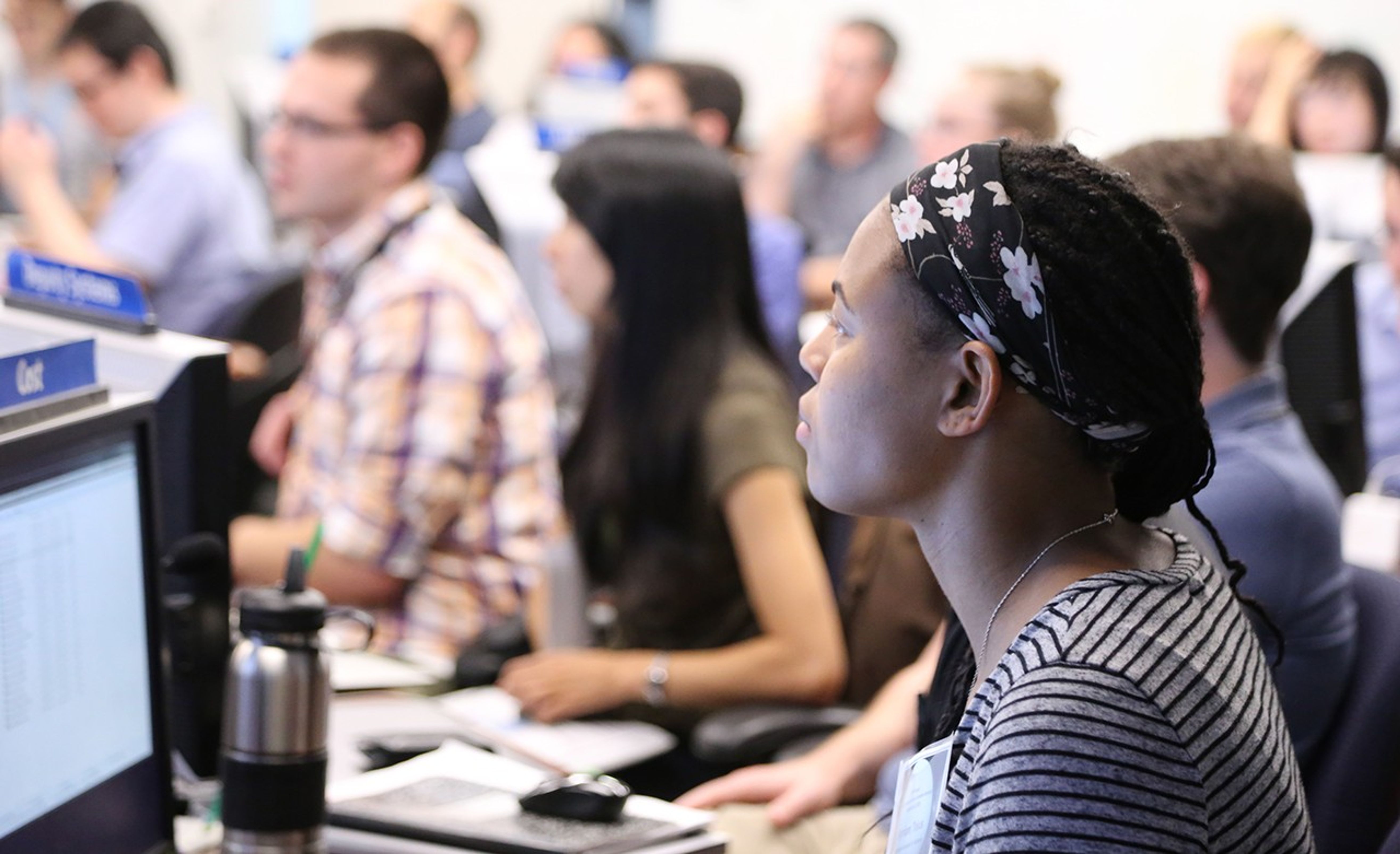 A woman sits in front of a computer console in a control room full of people. The sign on her computer says "Cost."