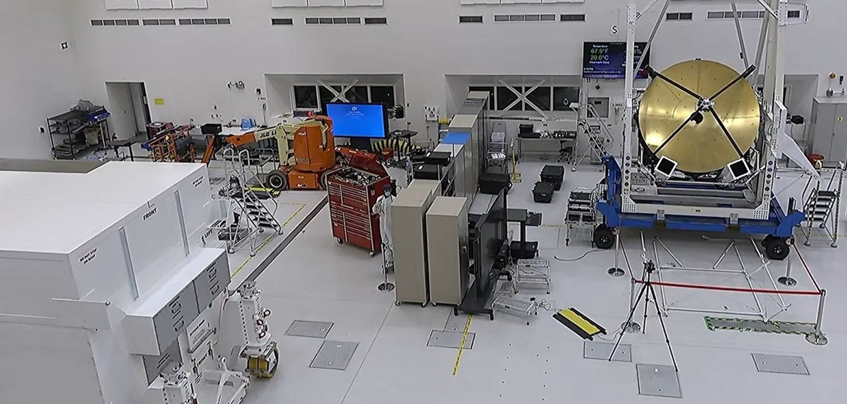 A large white room wiht lots of equipment where workers are building components of two spacecraft. To the left is a large white box. A red tool box in near the center along with a mannequin. To the right is a large gold dish.