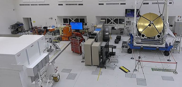 A large white room wiht lots of equipment where workers are building components of two spacecraft. To the left is a large white box. A red tool box in near the center along with a mannequin. To the right is a large gold dish.