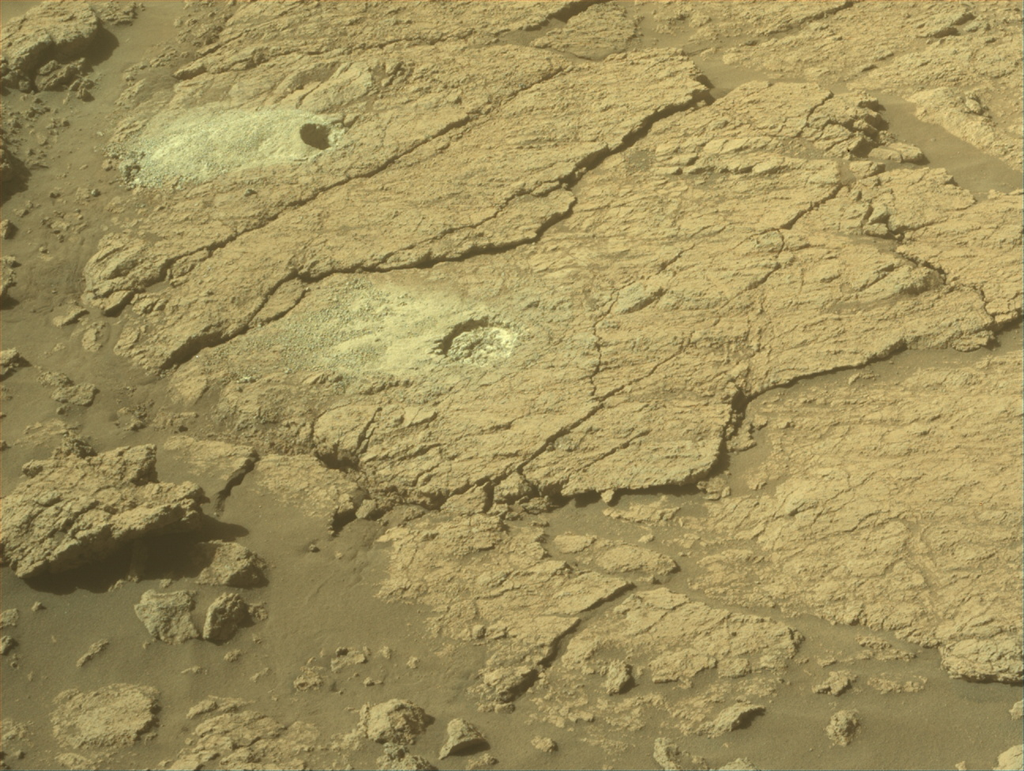 A circular drill hole with a pile of dust next to it on the surface of Mars.