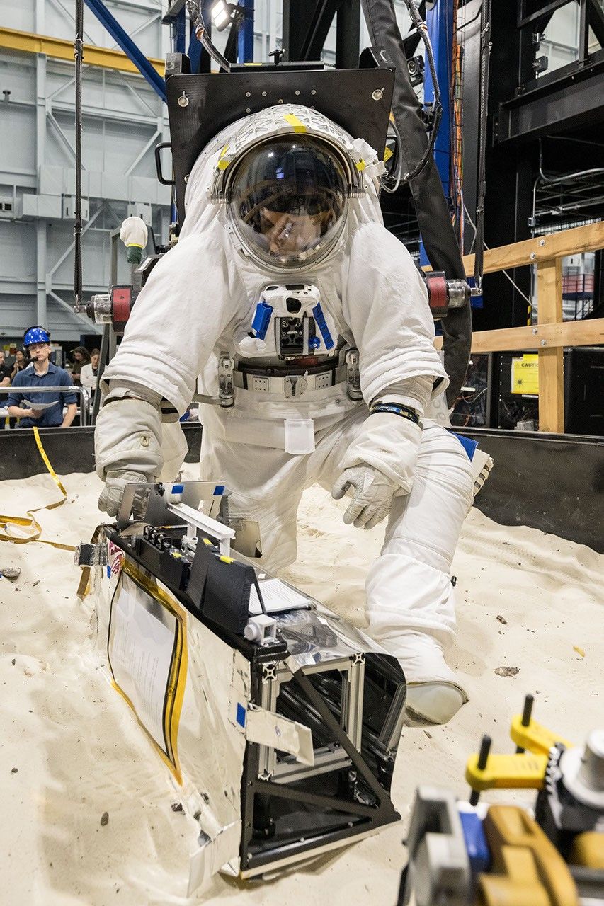 An astronaut in a white spacesuit kneels in simulated lunar soil while working with scientific equipment in a large testing facility. Staff members and support structures are visible in the… | Source: NASA Science