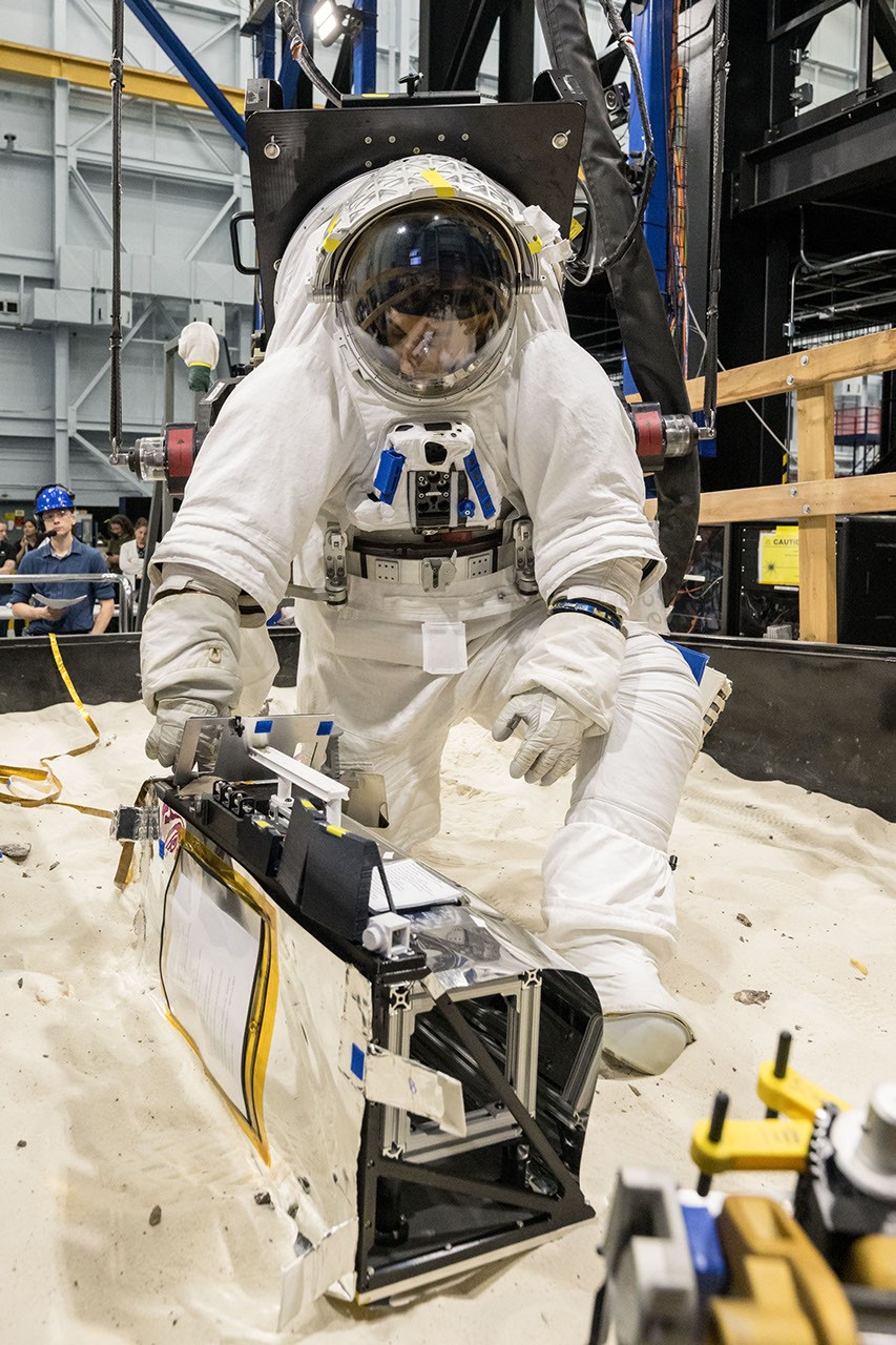 An astronaut in a white spacesuit kneels in simulated lunar soil while working with scientific equipment in a large testing facility. Staff members and support structures are visible in the background.