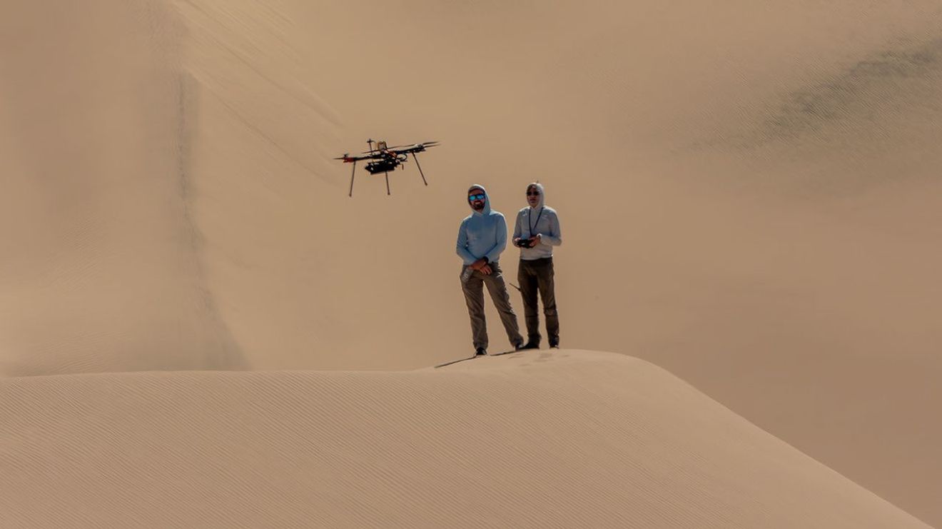 Two people wearing sun-protective gear stand on a sand dune, with a massive wall of sand filling the frame behind them. A drone hovers over their heads.