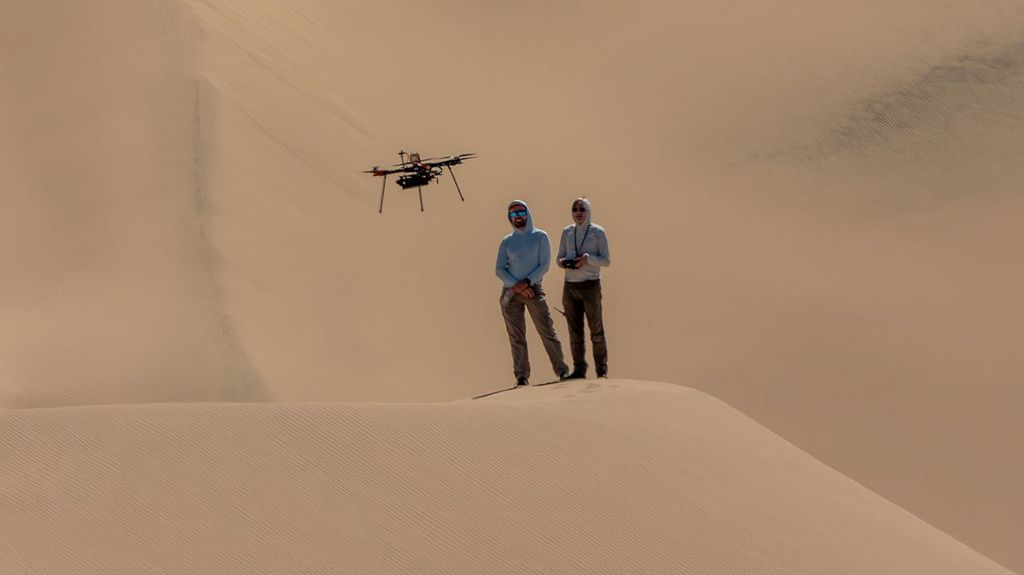 Two people wearing sun-protective gear stand on a sand dune, with a massive wall of sand filling the frame behind them. A drone hovers over their heads.