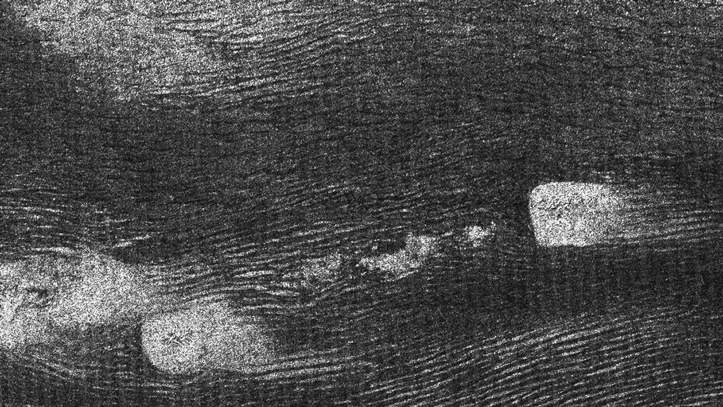 Grayscale image of a vast field of sand dunes, seen from orbit.  The swirling ridges of the dunes look almost like fingerprint patterns. A few blotches of dense white speckles interrupt an otherwise gray-to-dark-gray pattern.